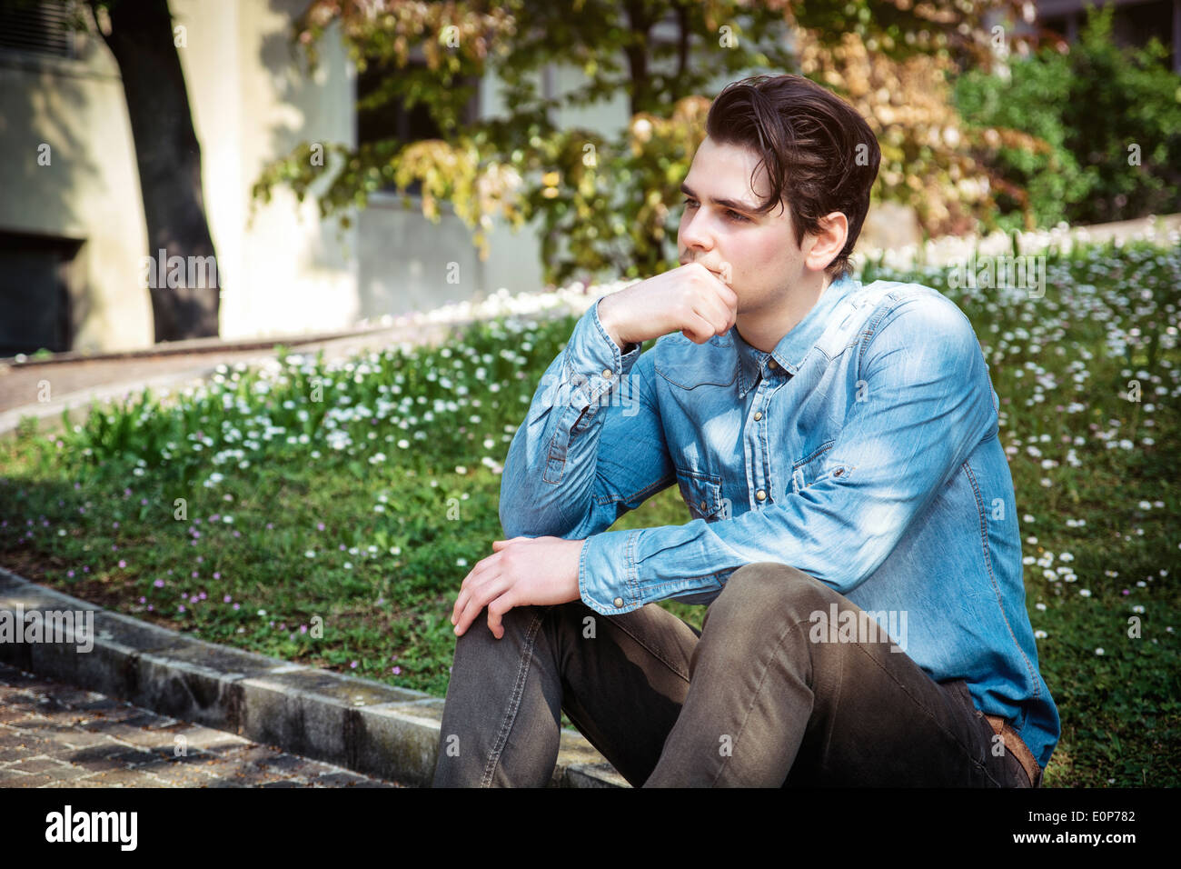 Junger Mann sitzt im Freien im öffentlichen Park, mit der Hand auf seinem Kinn Weg zu denken Stockfoto