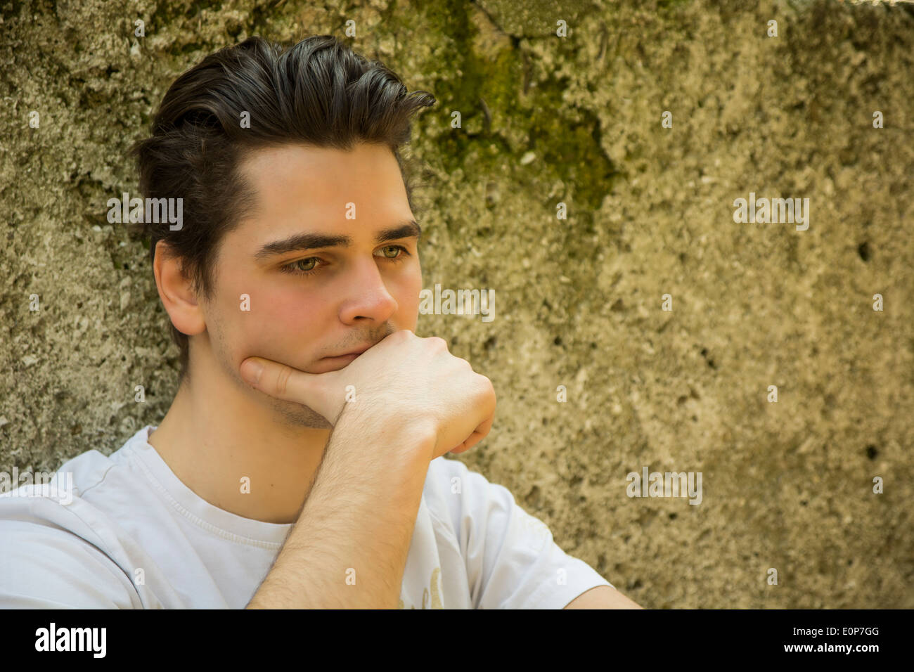Trauriger junger Mann mit Hand am Kinn denken, sitzen gegen Steinmauer Stockfoto