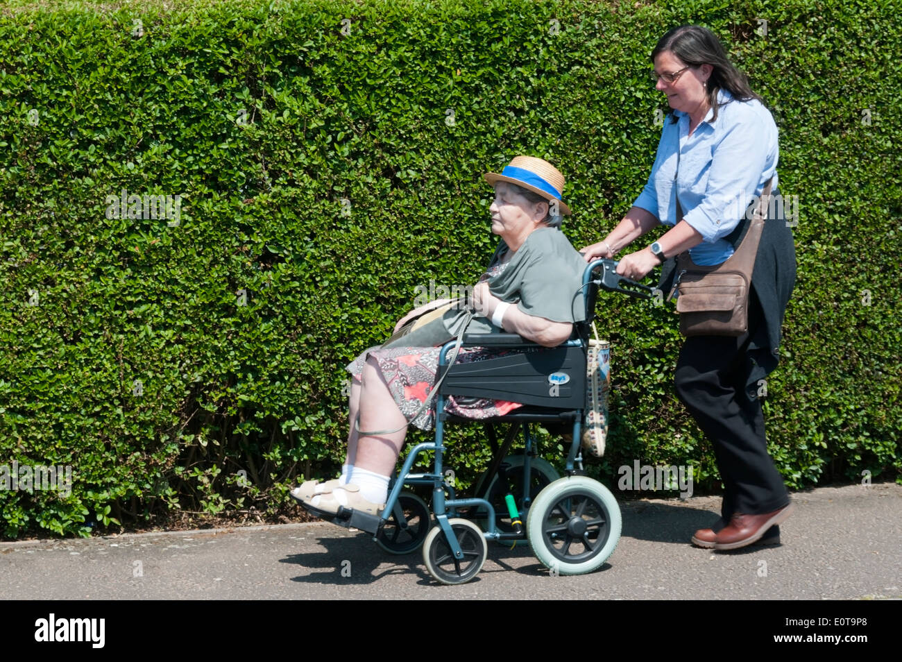 Eine ältere Frau trägt ein sonnenhut ist auf dem Gehsteig in einem Rollstuhl durch ihre Pfleger oder Assistent an einem sonnigen Tag geschoben. Stockfoto