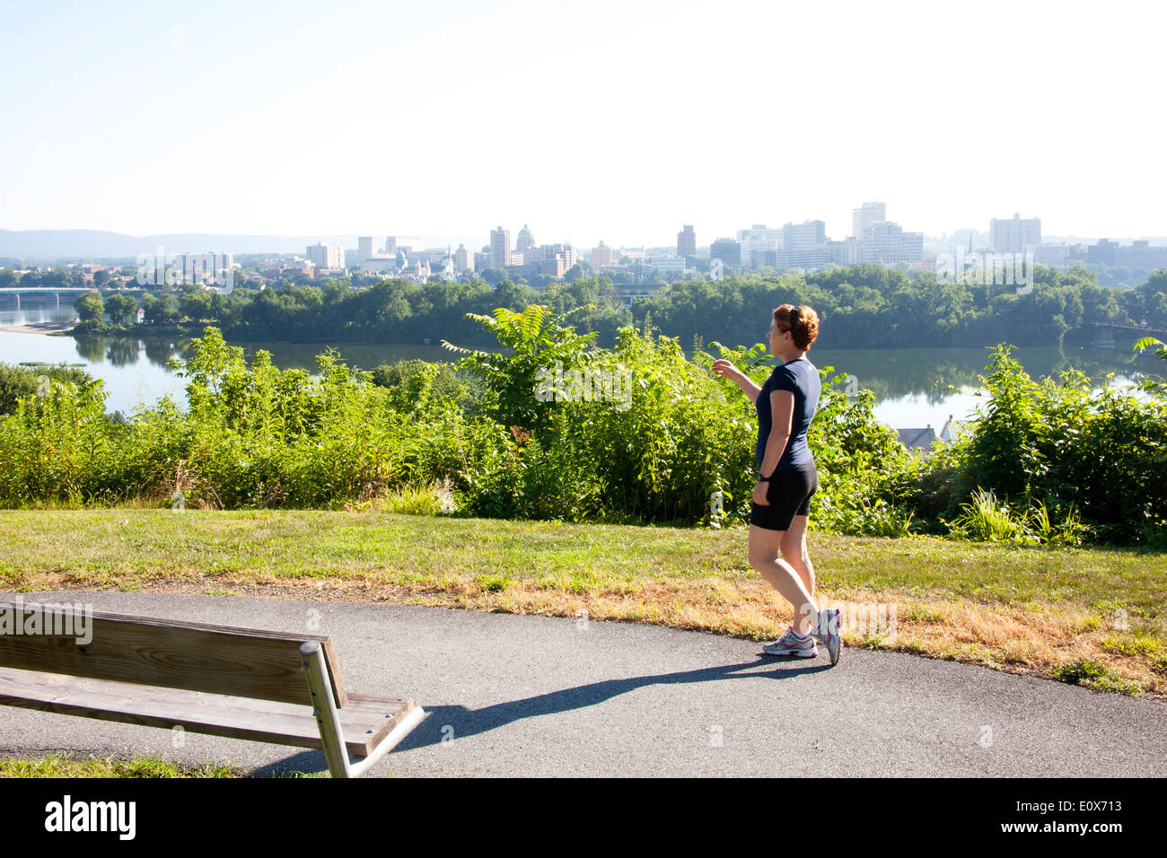 Weibliche Kraft zu Fuß entlang eines Pfads mit Blick auf einen Fluss Blick Skyline der Stadt an einem dunstigen, heißen Sommertag. Stockfoto