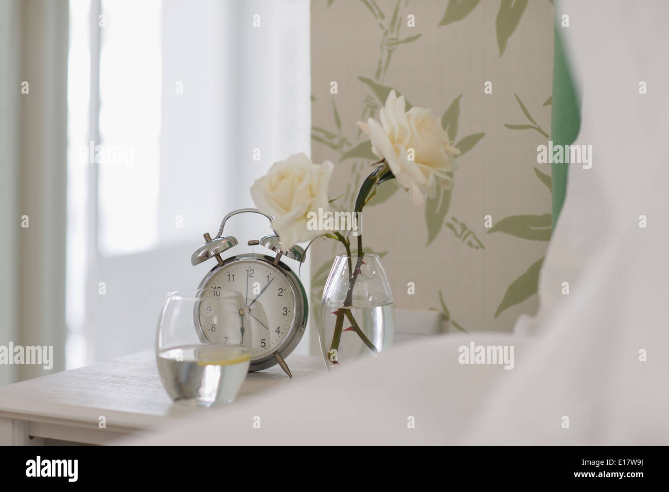 Weiße Rosen, Wecker und Wasserglas auf dem Nachttisch Stockfoto