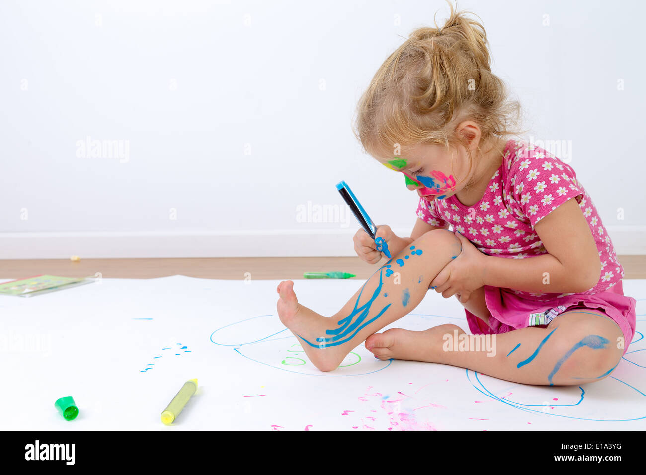 Niedlichen Kleinkind konzentriert ihre Beine vorsichtig mit bunten Stiften malen, nachdem sie ihr Gesicht gemalt Stockfoto
