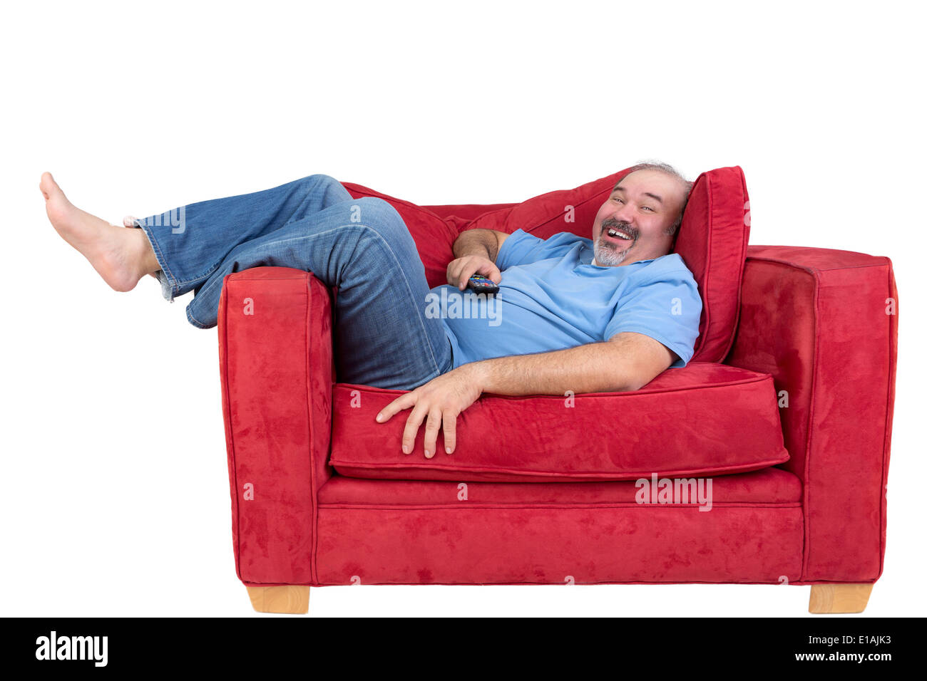 Aufgeregt Mann beobachtet Fernsehen lachend, als er wieder barfuß auf einem bequemen roten Sofa mit der Fernbedienung in der Hand liegt Stockfoto