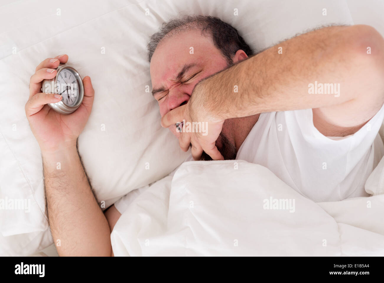 Mann im Bett zu gähnen, als er versucht, mit seinem in seiner Hand hielt Wecker aufwachen liegen Stockfoto