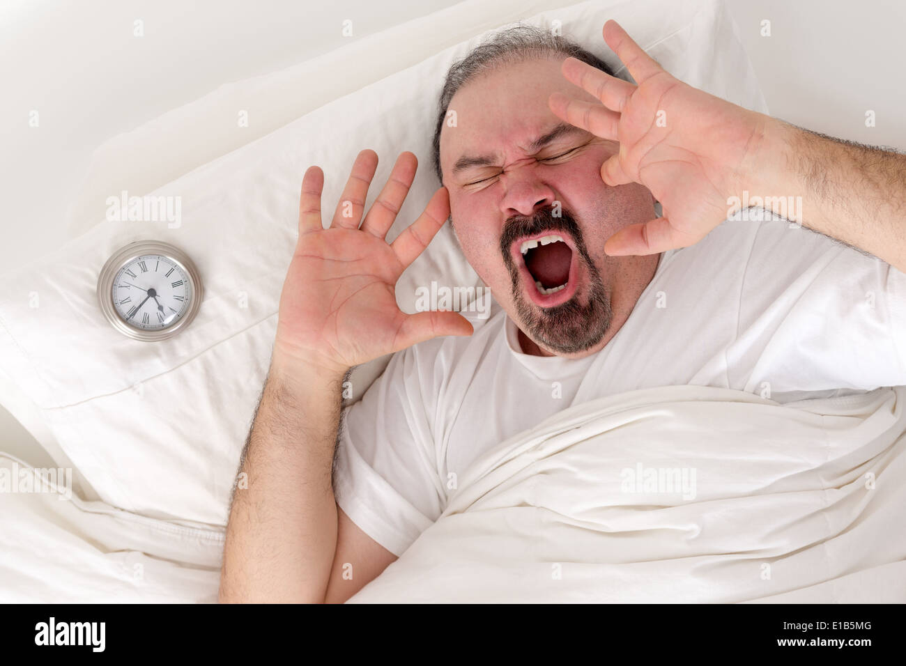 Im mittleren Alter bärtiger Mann Gähnen laut Ruhe im Bett mit seinen Wecker auf dem Kissen neben ihm, als er versucht, nach dem Aufwachen Stockfoto