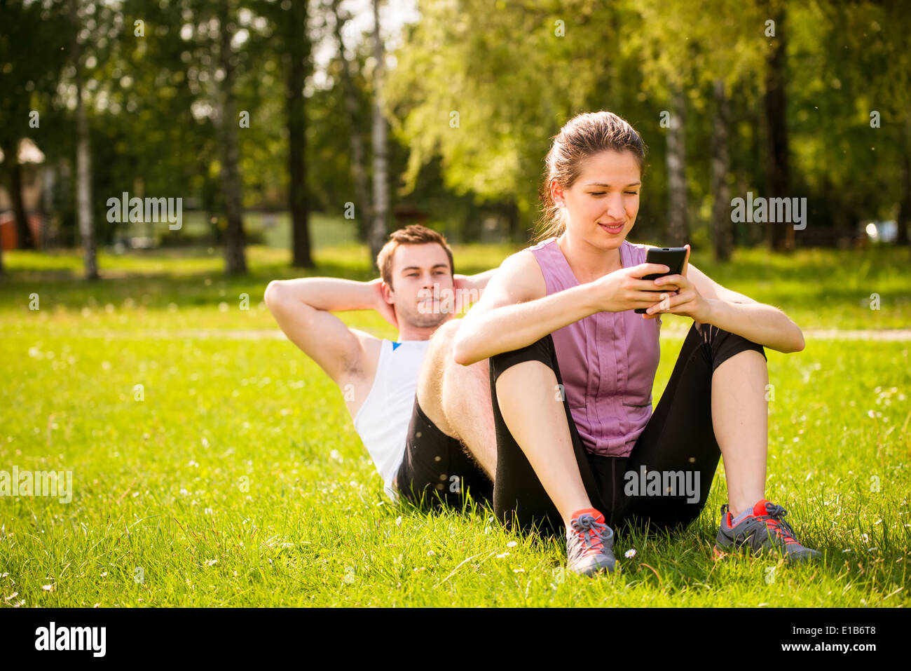 Mann macht Sit-ups, während Frau sitzt auf seinen Füßen und spielen mit Handy Stockfoto