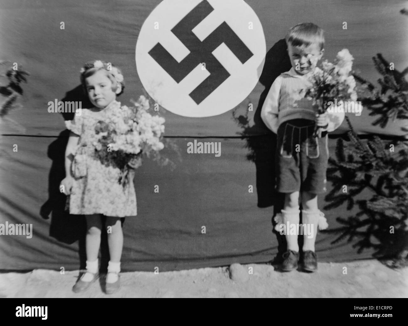 Junge deutsche Mädchen und jungen im Rahmen einer Nazi-Zeremonie, ca. 1937. Sie halten Blumen und stehen neben einem großen Hakenkreuz in Brandl bin Stockfoto