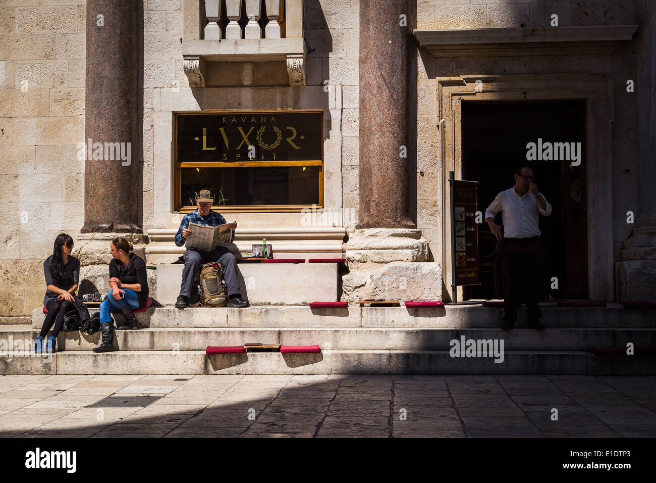 Zwei Frauen sprechen, während ein Mann eine Zeitung am Peristil im Diokletianpalast, Split, Kroatien liest. ein Kellner blickt auf Stockfoto