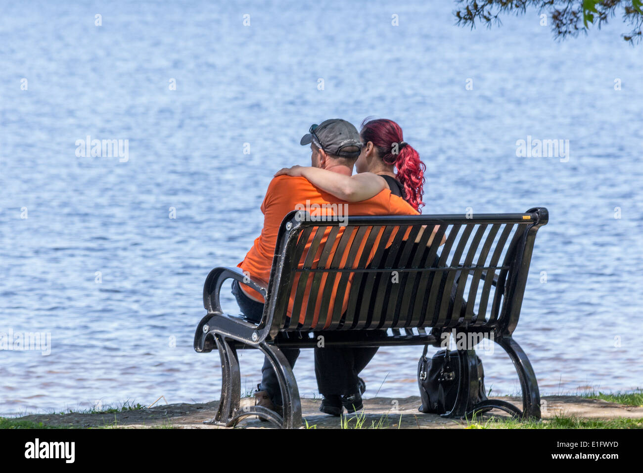 Junges Paar mit Arme umeinander auf Parkbank mit Blick auf den See. Stockfoto