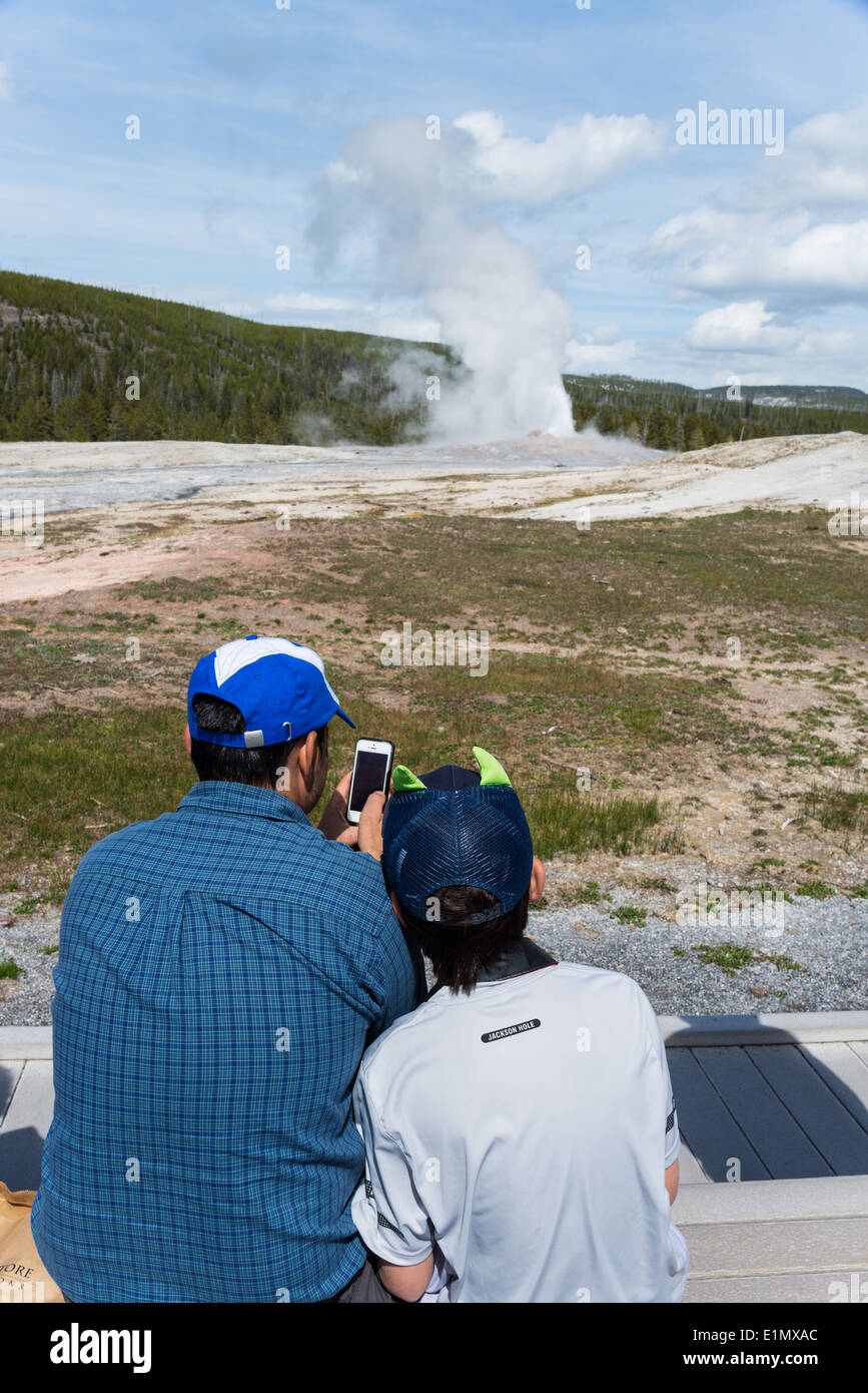 Vater und Sohn machen Handy Fotos von Old Faithful Geysir Ausbruch. Yellowstone-Nationalpark, Wyoming, USA. Stockfoto
