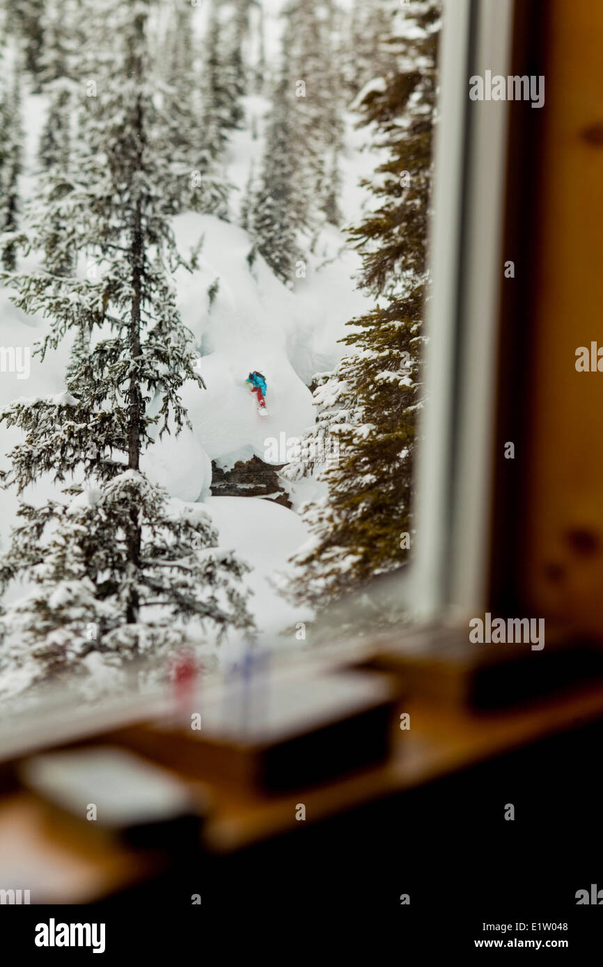 Backcountry Snowboarder fällt eine Klippe während eingerahmt durch ein Lodge-Fenster, Eisfall Lodge, Golden, BC Stockfoto