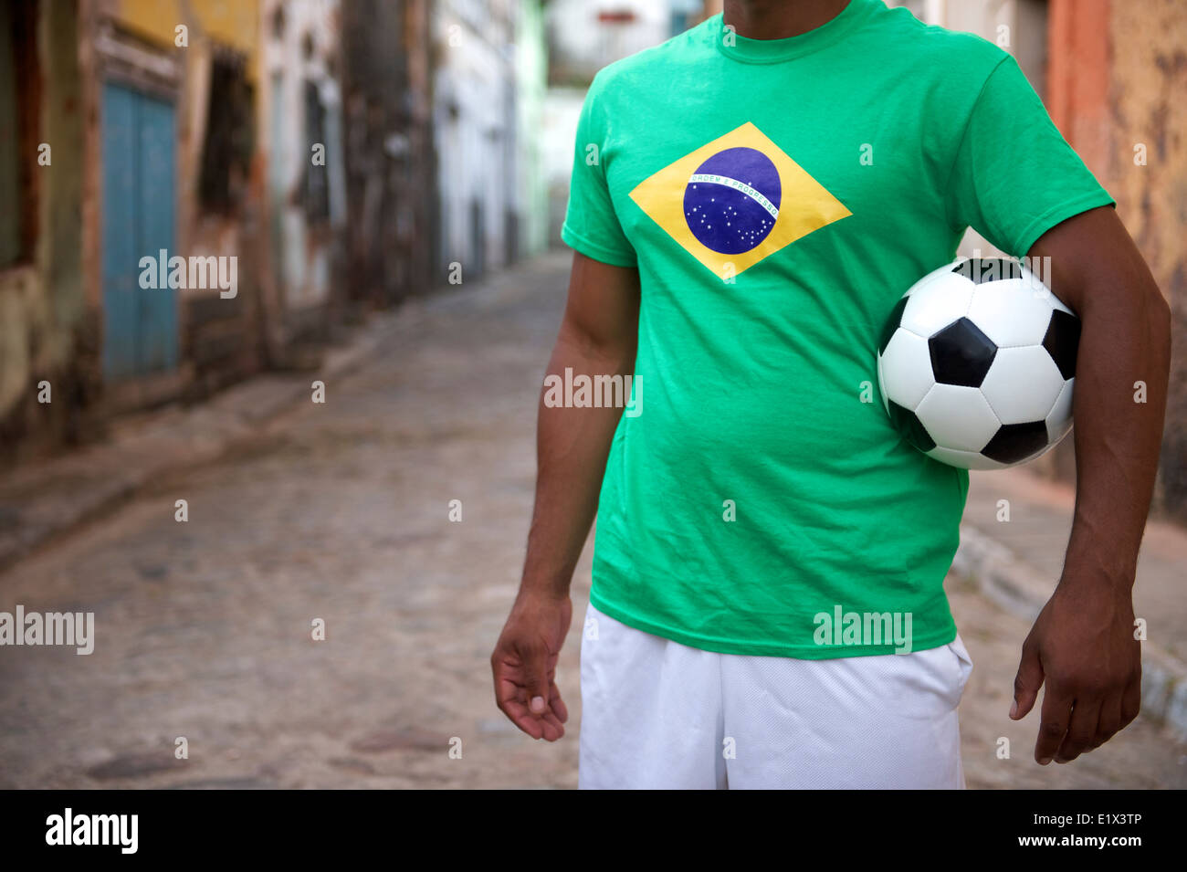 Brasilianische Street-Football-Spieler stehen in Brasilien Flagge T-shirt Holding Fußball ball in einem alten rustikalen Dorf Stockfoto