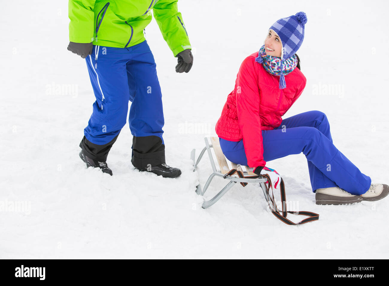 Glückliche junge Frau auf Schlitten sitzend und Blick auf den Mann im Schnee Stockfoto