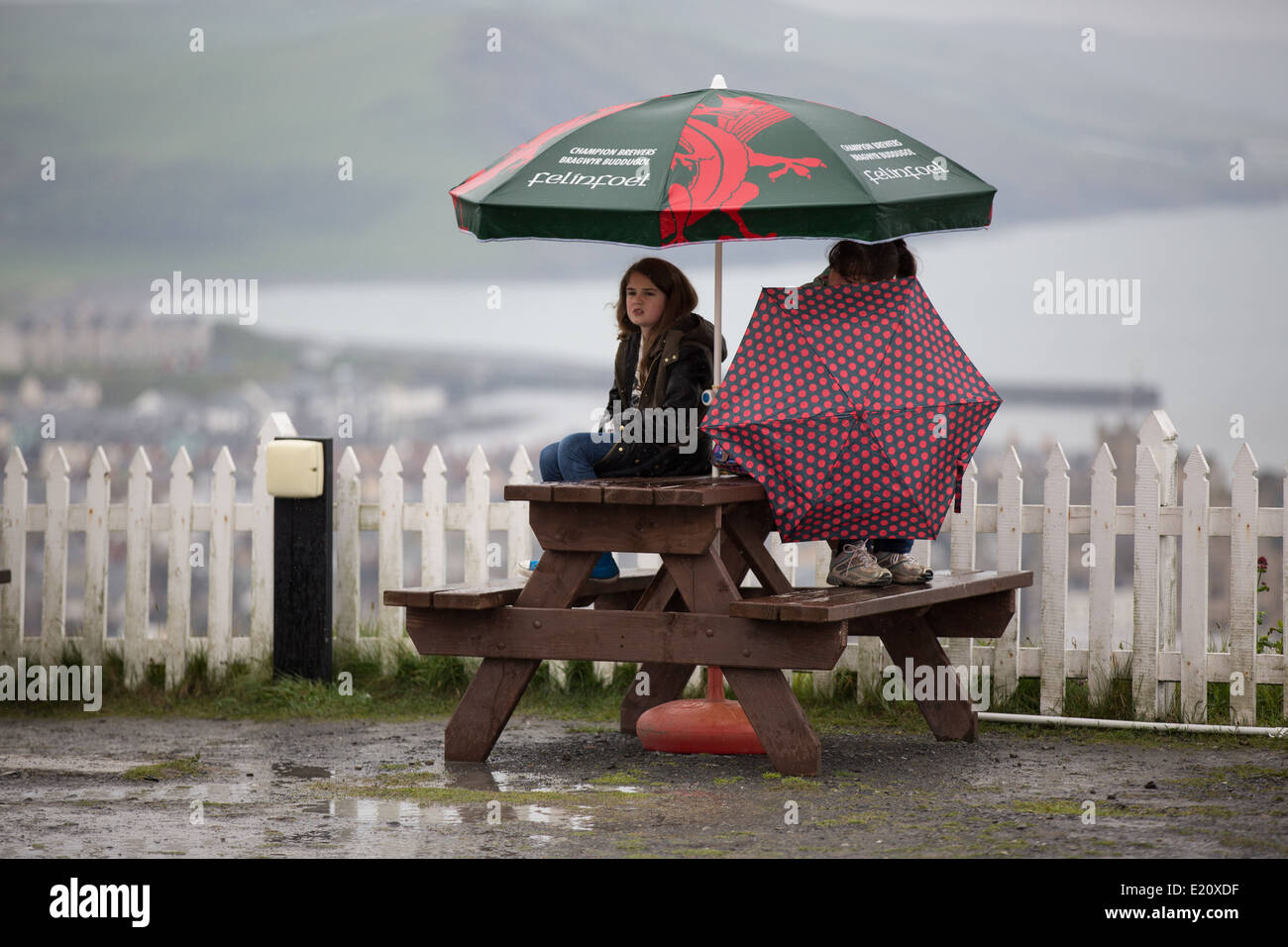 Zwei Mädchen im Teenageralter an einer Picknickbank im Regenschutz unter Sonnenschirmen im Regen sitzen. Stockfoto
