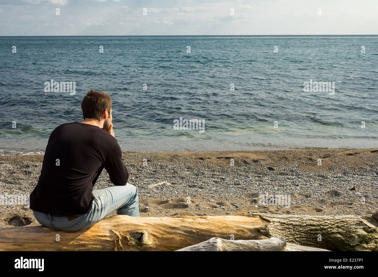 Mann sitzt auf einem Baumstamm, starrt auf das Meer Stockfoto