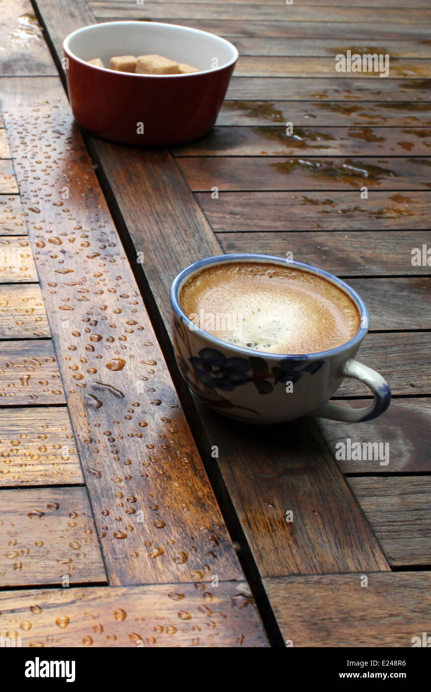 Tasse Kaffee mit Schaum und eine Schüssel mit braunem Zuckerwürfel auf einem Holztisch mit Regen fällt auf ihn. Stockfoto