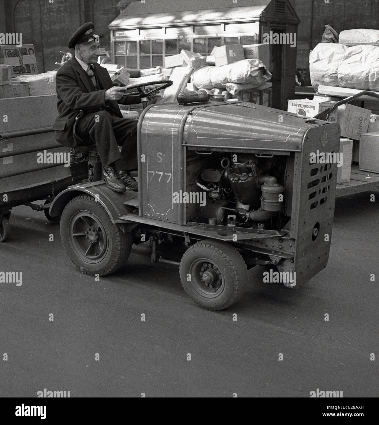 1950, historische, eine uniformierte Briitsh Eisenbahnen Porter auf dem Zusammentreffen in der Londoner Waterloo Bahnhof auf einer kleinen motorisierten Fahrzeug Ziehen eines Anhängers Verschieben von Paketen. Stockfoto
