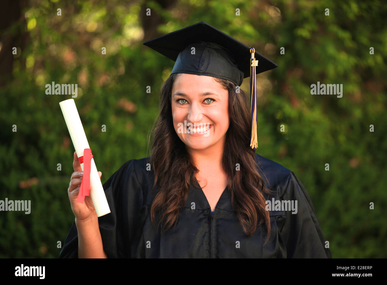 Attraktives Mädchen mit blauen Augen und braunen Haaren trägt eine Graduierung Mütze und Mantel und hält ein Diplom. Stockfoto