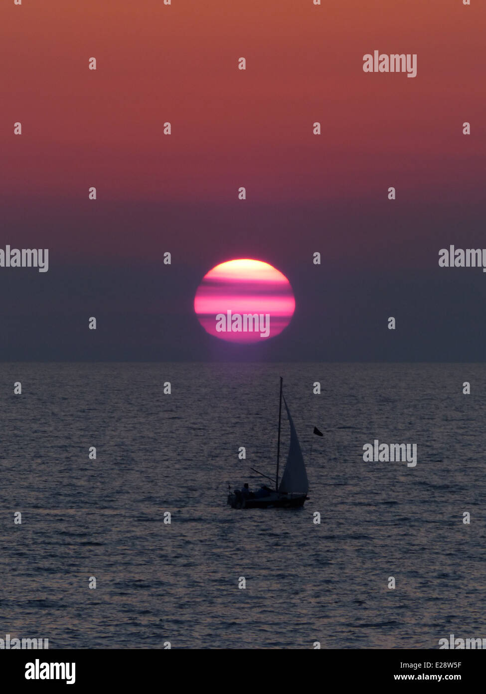 Eine Silhouette eines kleinen Segelboots in der Dämmerung mit einem großen roten und rosa gestreifte Sonne im Hintergrund Stockfoto