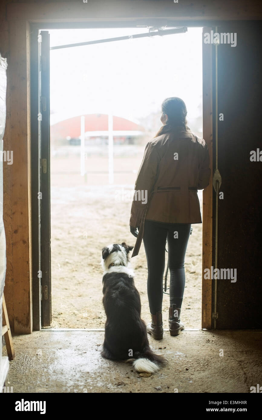 Voller Länge Rückansicht der jungen Frau mit Hund gelehnt in Tür Pferdestall Stockfoto