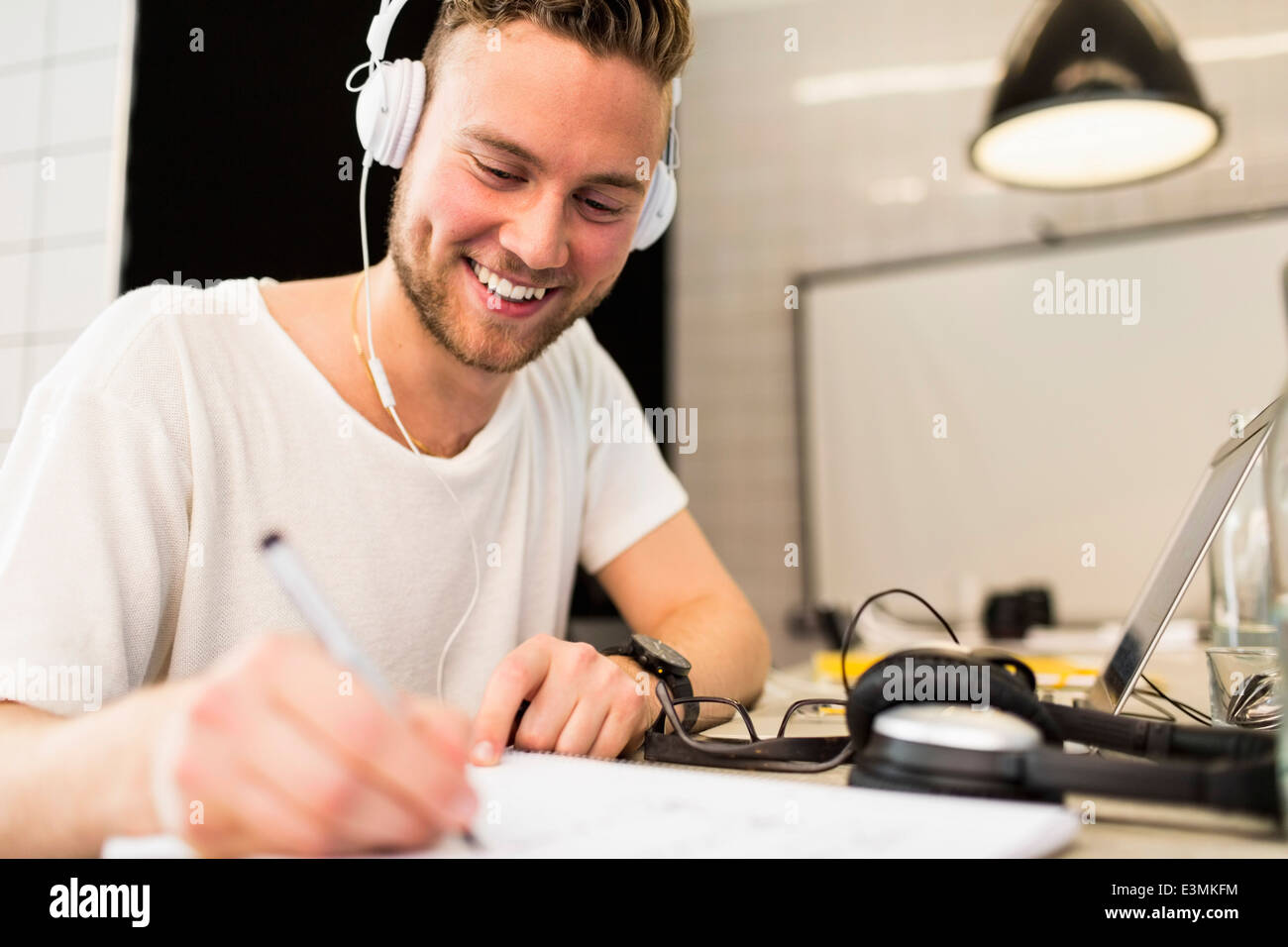 Glücklich junger Geschäftsmann mit Kopfhörern während des Schreibens in Buch am Kreativbüro Stockfoto