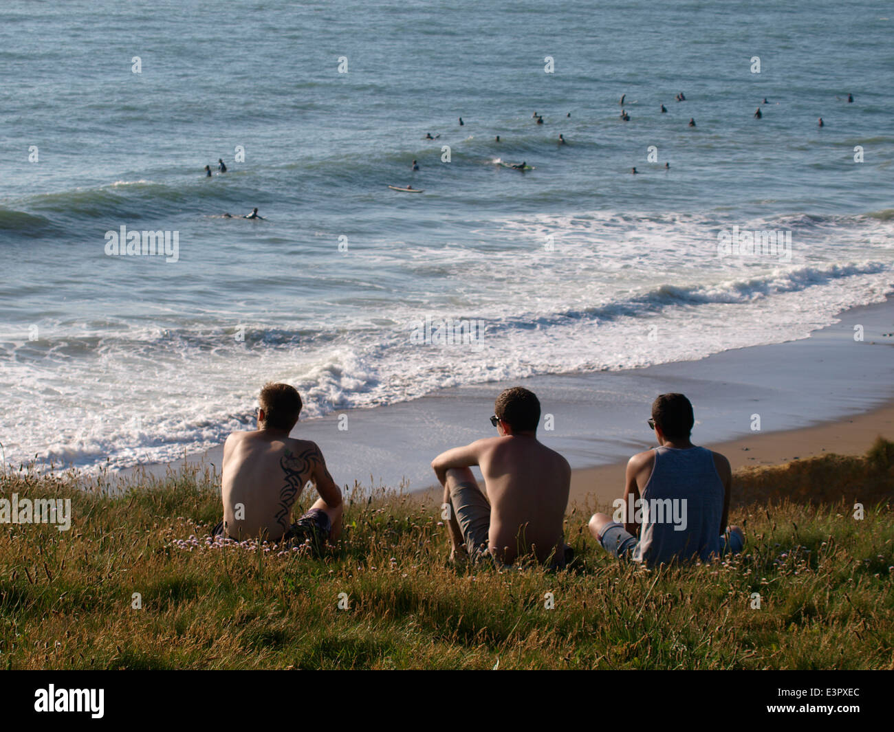 Drei junge Männer saßen auf einer Klippe mit Blick auf das Meer, Widemouth Bay, Bude, Cornwall, UK Stockfoto