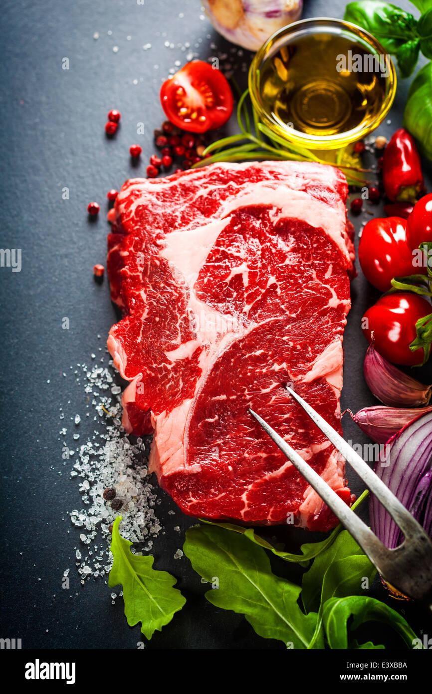 Rohes Rindfleischsteak mit Fleischgabel und Zutaten auf einem dunklen Hintergrund Schiefer Stockfoto