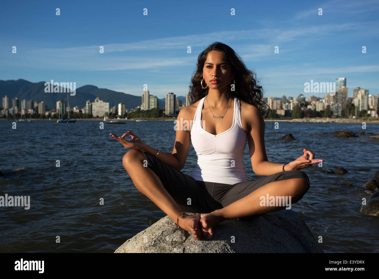 Eine junge Indianerin ist auf einem Felsen mit den Ozean und die Innenstadt von Vancouver im Hintergrund meditieren. Stockfoto