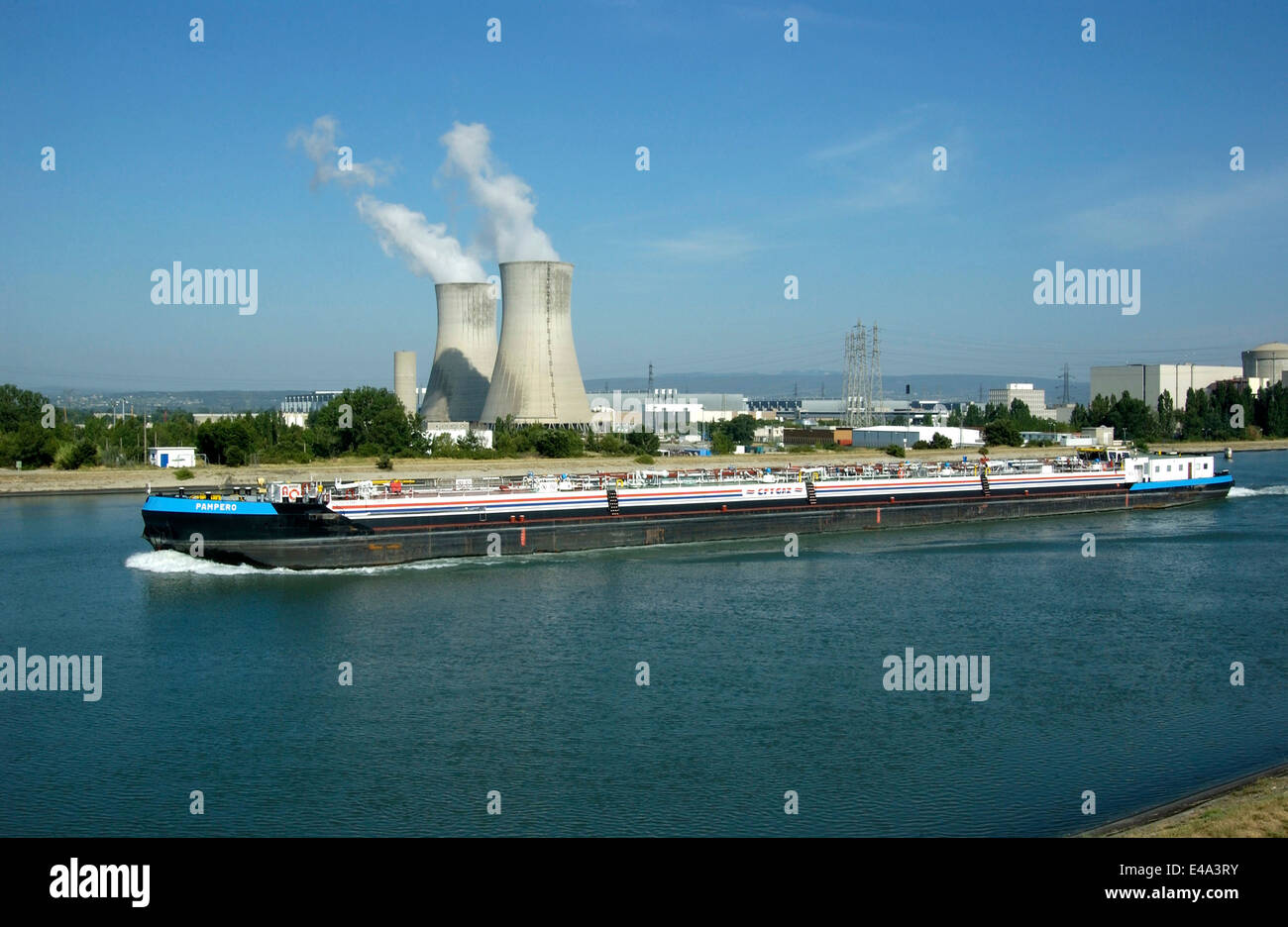 Ein Binnenschiff fährt über die Rhone und passiert das Kernkraftwerk Tricastin in Drôme, Frankreich. Dampf steigt aus Kühltürmen unter einem hellen Blau auf Stockfoto