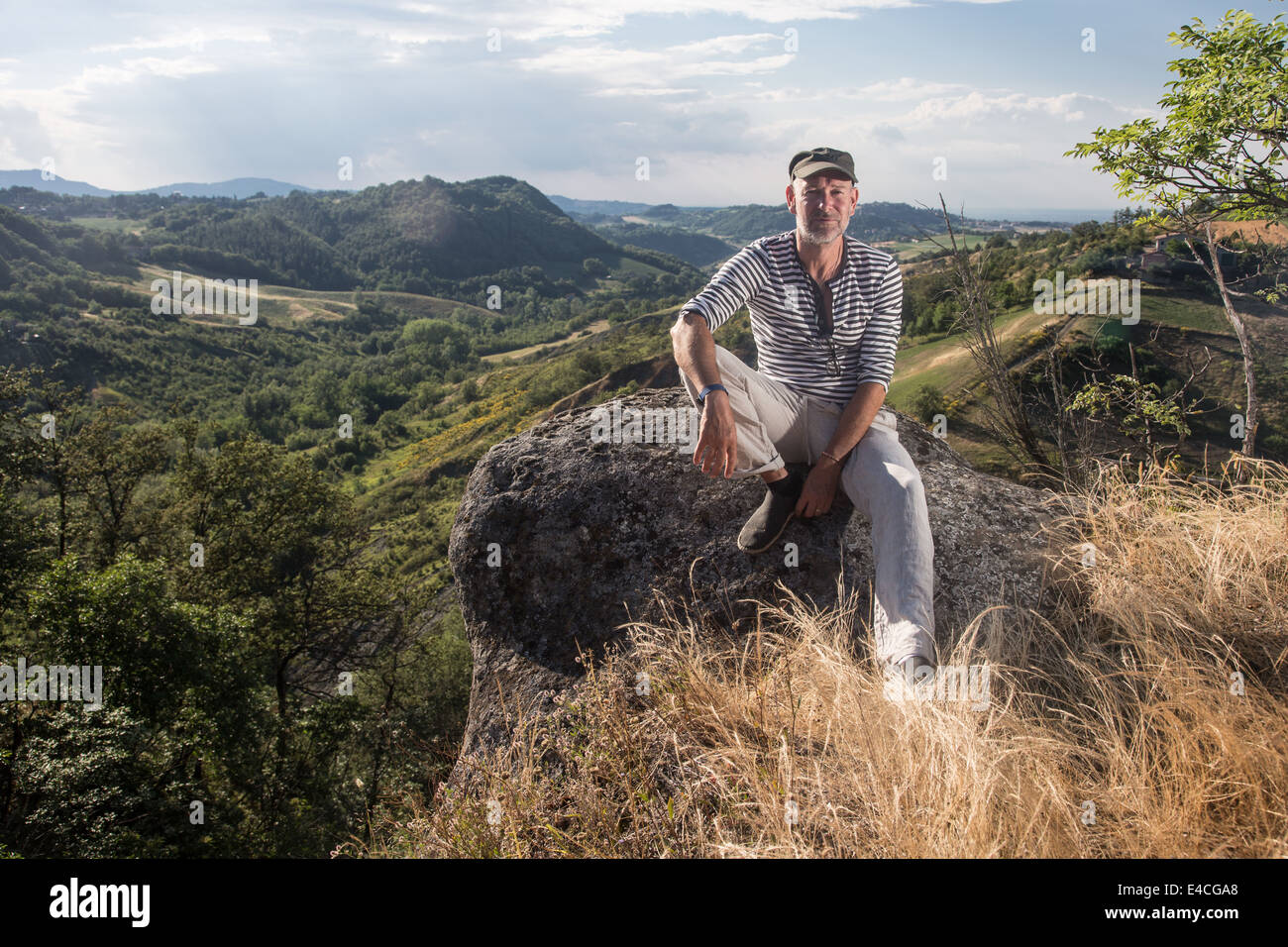 ernster Mann sitzt auf Felsen tragen gestreifte Spitze und Kappe in Landschaft Blick direkt in die Kamera Stockfoto