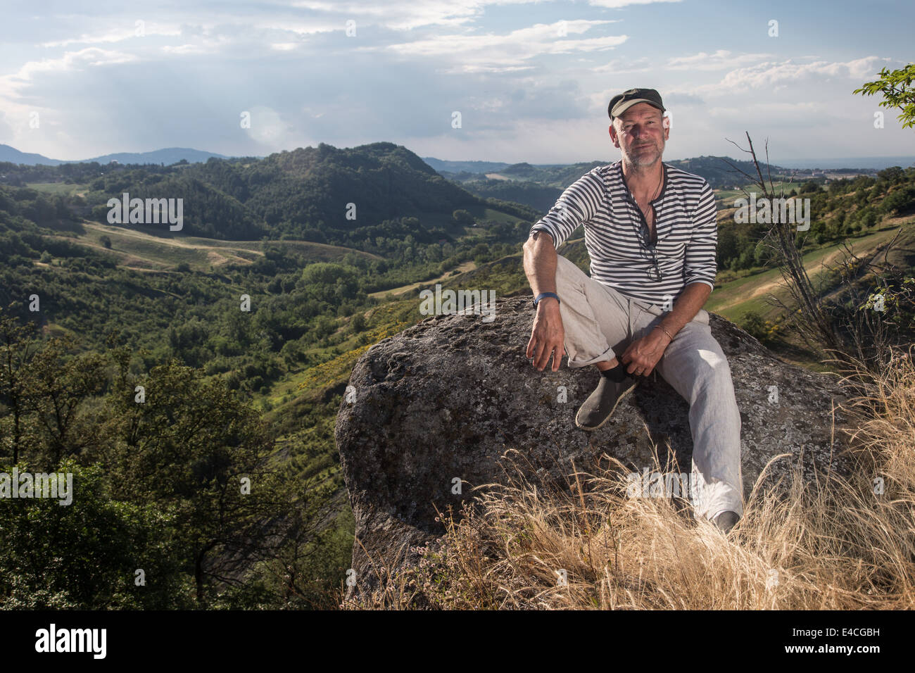 ernster Mann sitzt auf Felsen tragen gestreifte Spitze und Kappe in Landschaft Blick direkt in die Kamera Stockfoto
