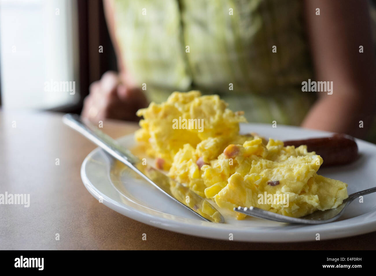 Rührei mit Frühstückswurst auf einem weißen Teller mit einer Frau, die im Hintergrund auf einem Tisch zu sitzen. Stockfoto
