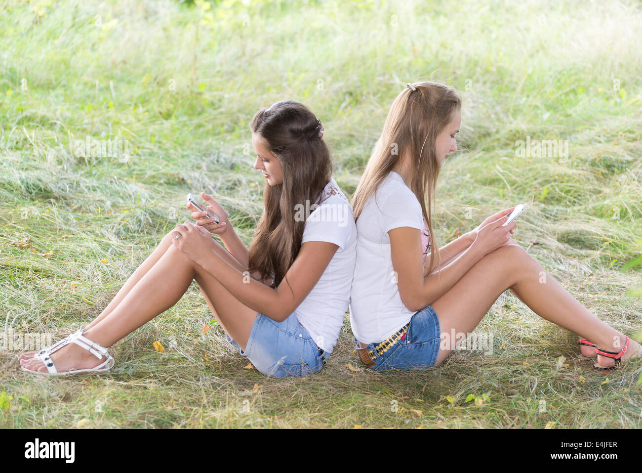 Zwei Mädchen im Teenageralter mit Handys sind auf dem Rasen sitzen. Stockfoto