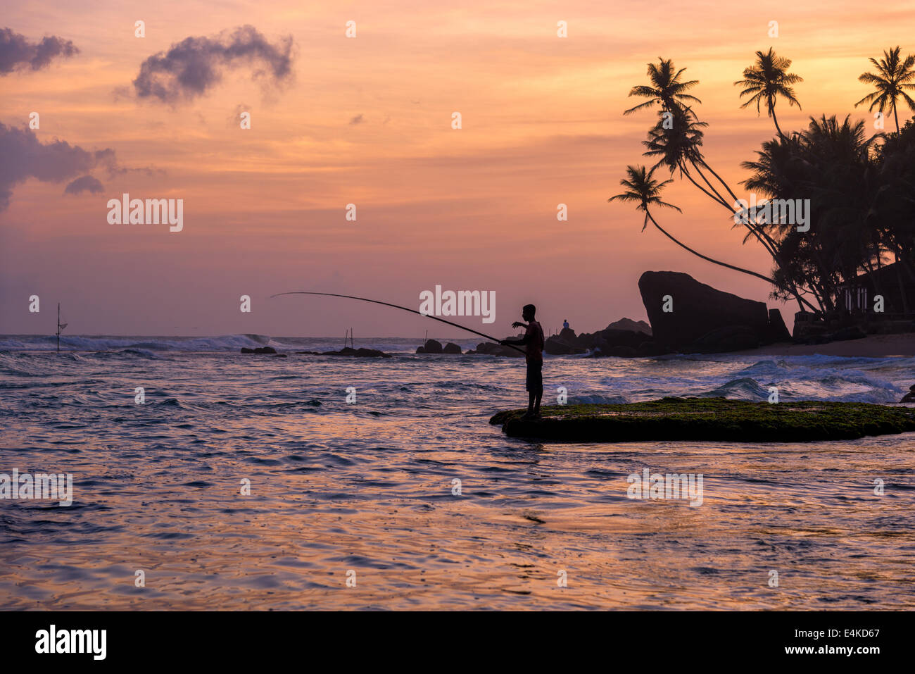 Ein Fischer, Palmen und Felsen in der Silhouette bei Sonnenuntergang an einem Strand bei Unawatuna auf der westlichen Küste von Sri Lanka Stockfoto