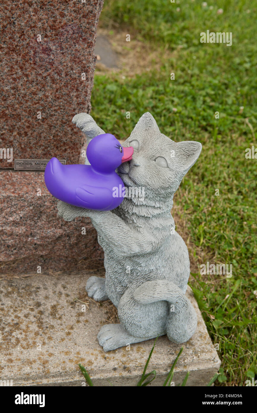 Konkrete Statue einer Katze in einer Grabstätte umarmt eine lila Gummiente. Stockfoto