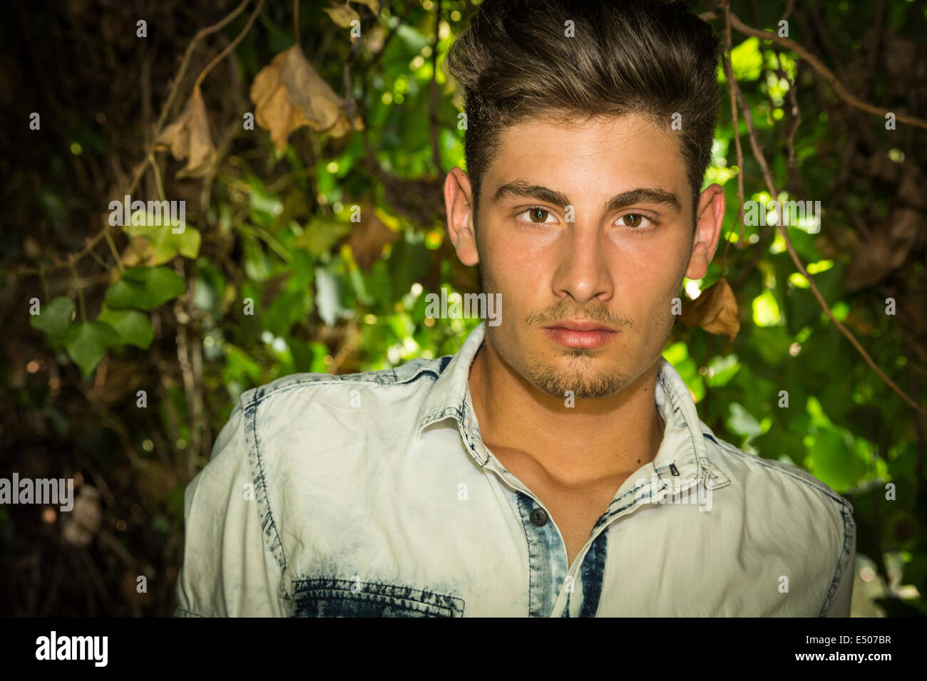 Hübscher junger Mann, Blick in die Kamera. Pflanzen und Sträucher um ihn herum Stockfoto