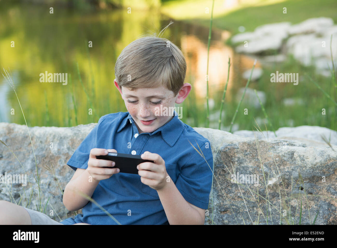 Ein kleiner Junge im Freien sitzen lehnte sich gegen einen Felsen mit einem handheld elektronische Spiel. Stockfoto