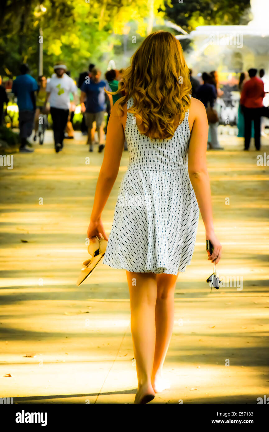 Rückansicht einer jungen weißen Hündin mit langen Haaren, die barfuß in einem Sundress in Richtung des Forsyth Park Springbrunnens bei Sonnenuntergang, Savannah, GA, USA, geht Stockfoto