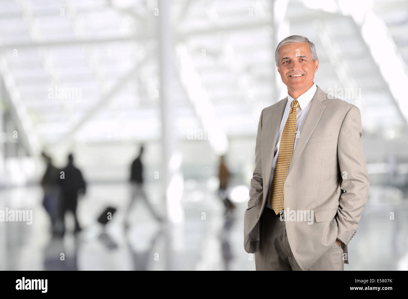 Reife Geschäftsmann in Flughafen Halle mit unscharfen Reisenden im Hintergrund. Man hat die Hände in den Hosentaschen und lächelt. Stockfoto
