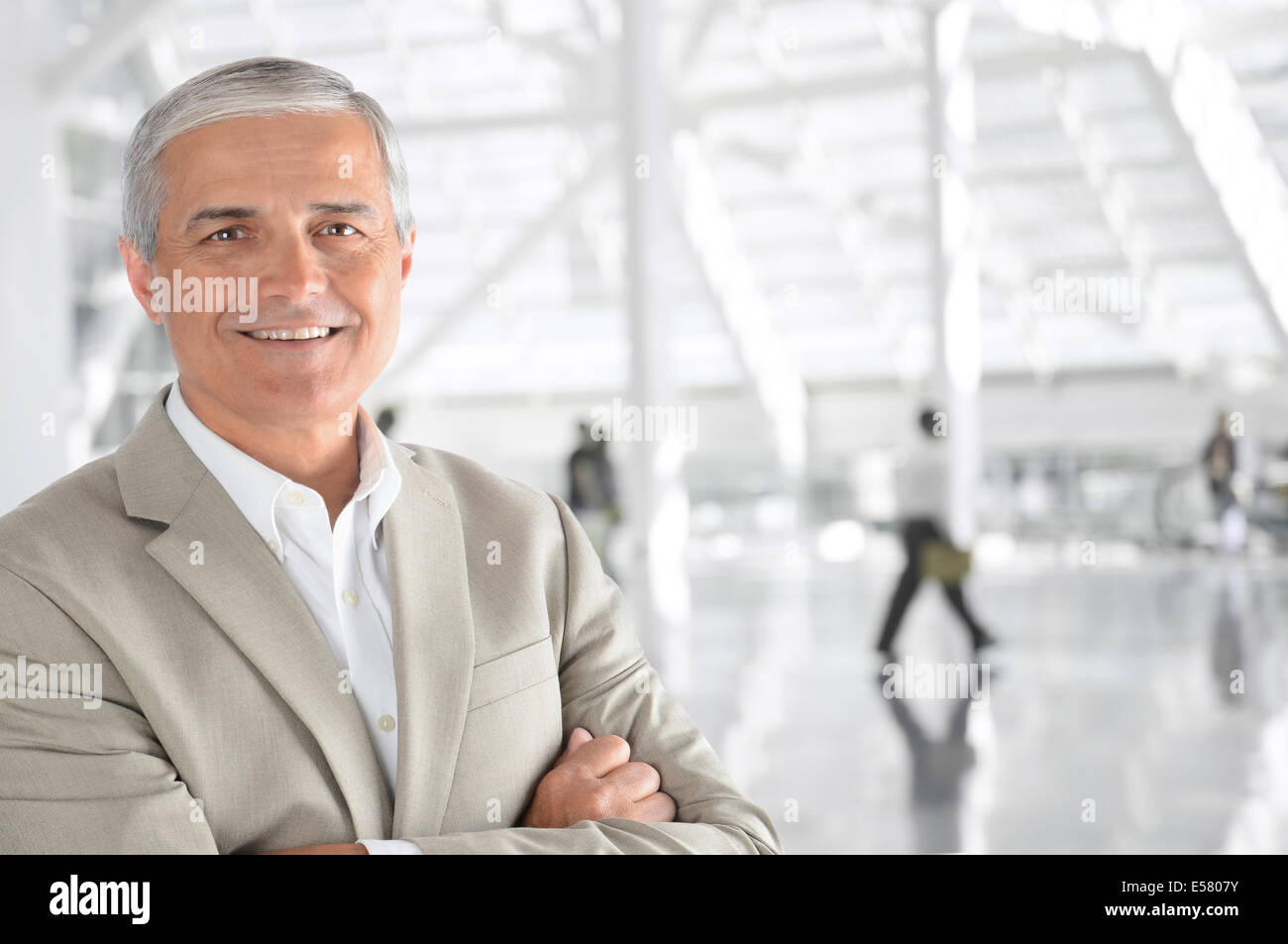 Nahaufnahme eines ausgereiften Geschäftsmann mit seinen Armen gefaltet in einer Flughafen-Halle. Der Mann lächelt in die Kamera mit unscharfen pass Stockfoto