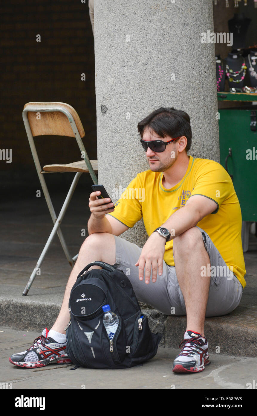 Junge Mann trägt ein gelbes Hemd und dunkle Sonnenbrille, SMS mit seinem Handy in Covent Garden in London Stockfoto