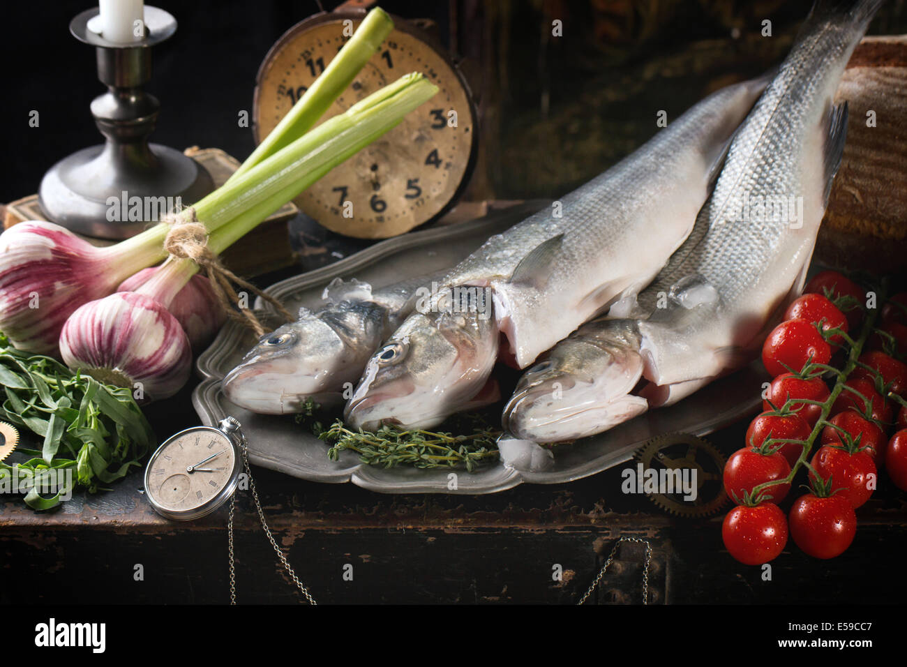 Stillleben mit rohem Fisch Seebarsch, Kräuter, Gemüse und Vintage-Uhr Stockfoto