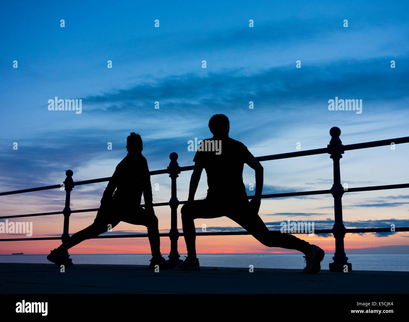 Jogginghose, Mann und Frau, die sich ausdehnt und sich bei Sonnenaufgang mit Blick auf das Meer erwärmt. GROSSBRITANNIEN Stockfoto