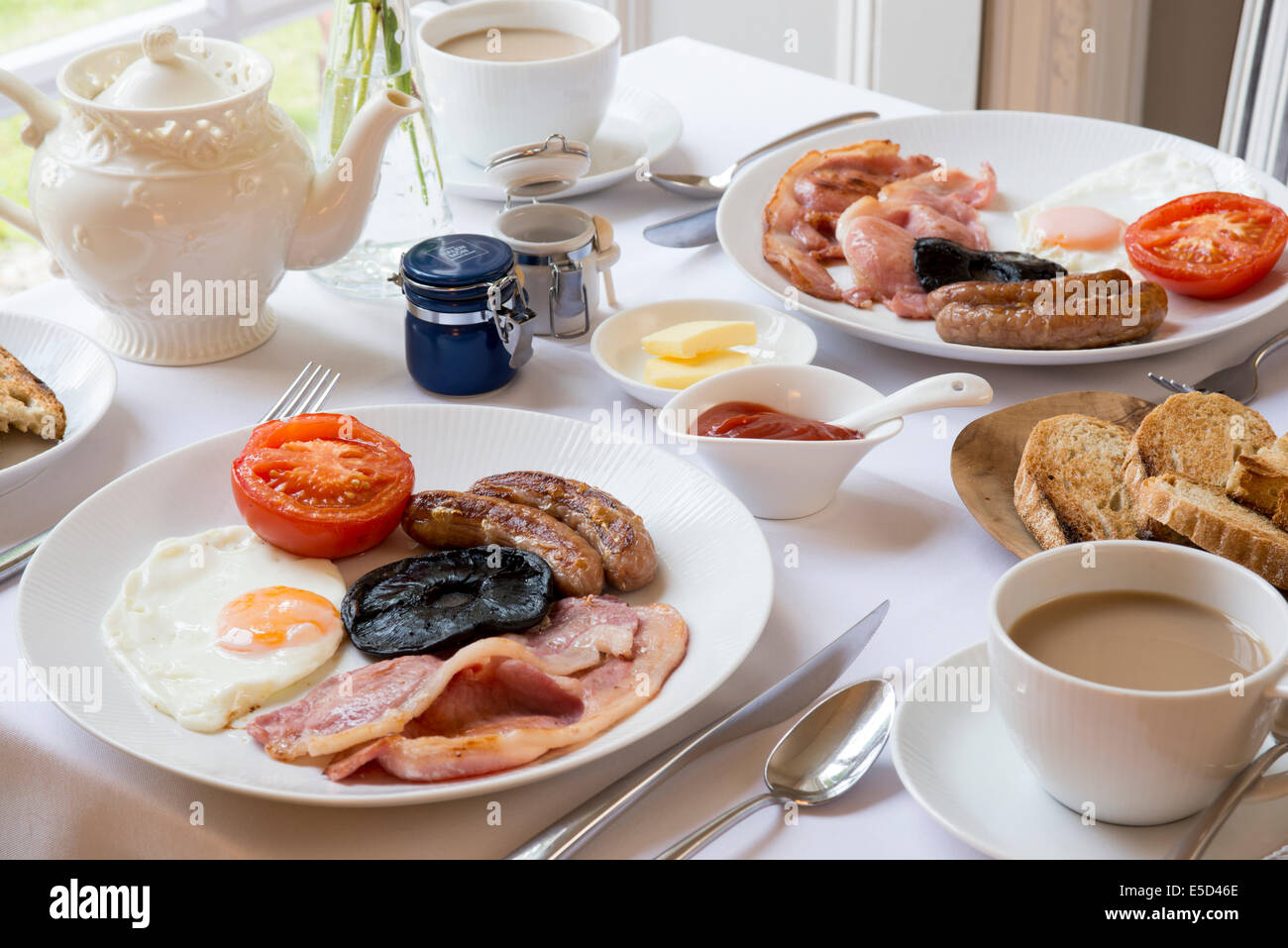 Traditionelles englisches Frühstückstisch ausgebreitet durch Fenster Innenraum mit Tee Toast Speck, Würstchen und Eiern auf Tischdecke Stockfoto