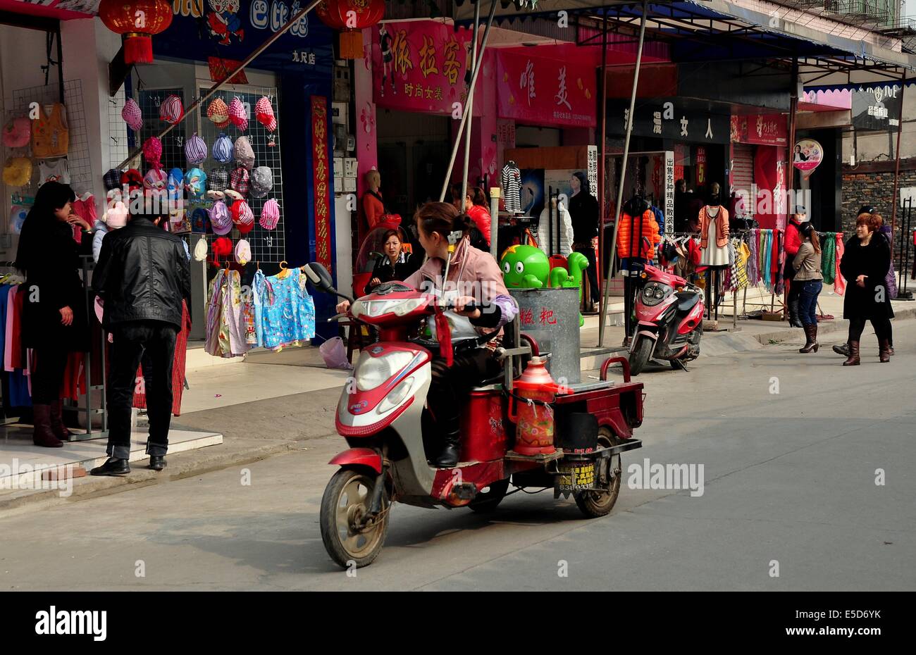 JIU-CHI-TOWN, CHINA: Frau reitet auf ihrem Motorrad Warenkorb Blicke auf ein Bekleidungsgeschäft an einer belebten Einkaufsstraße Stockfoto