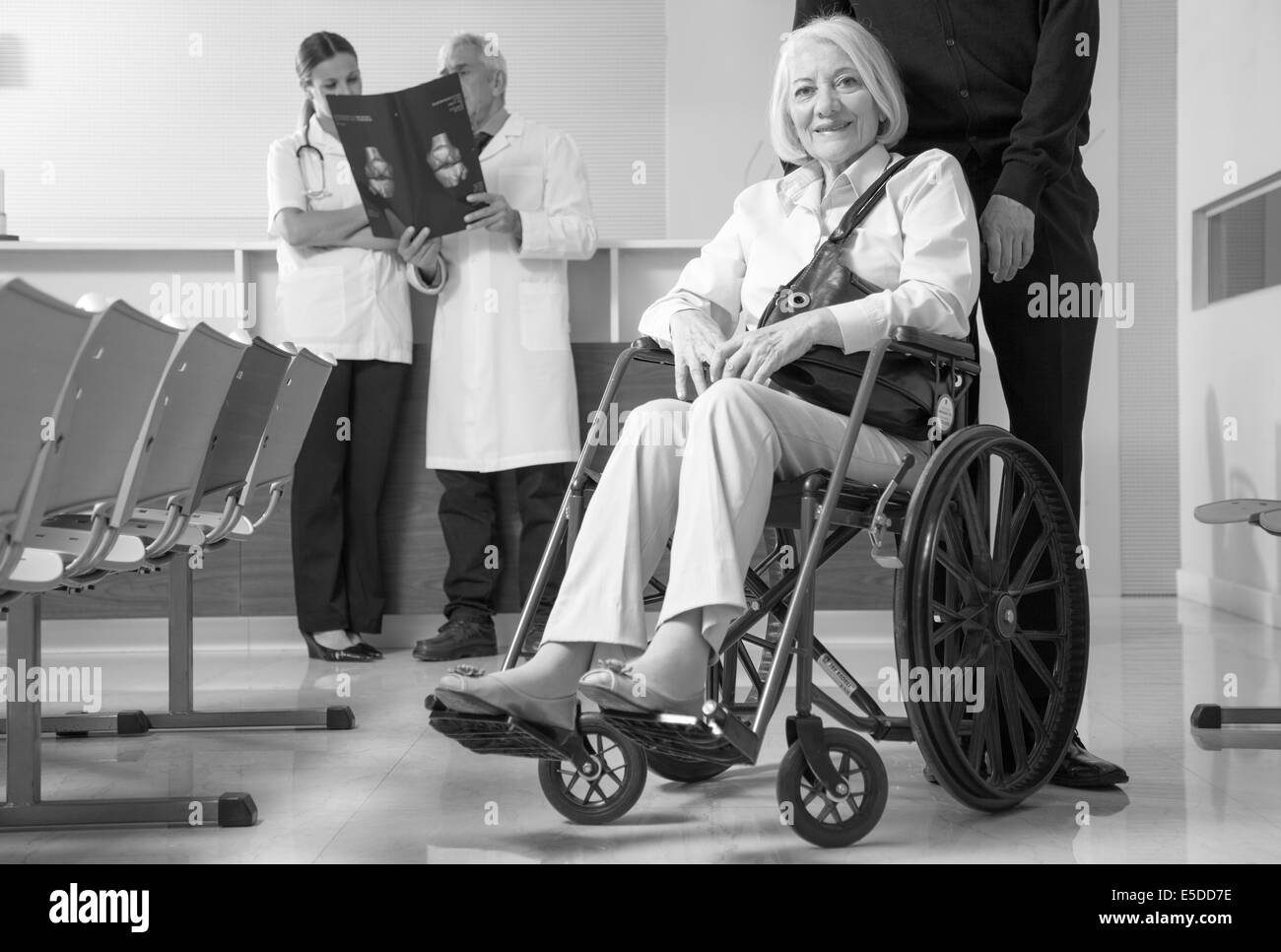 Reife Frau im Rollstuhl mit ihrem Ehemann im Krankenhaus. Ärzte analysieren Scan im Hintergrund. Stockfoto