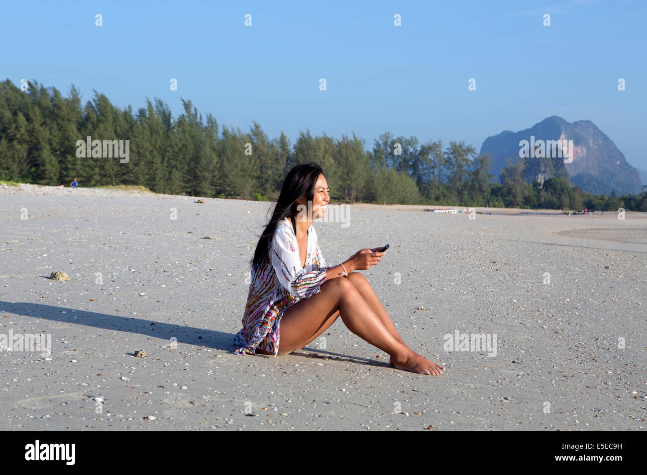 Eine einzelne junge asiatische Frau lächelnd, als sie eine SMS-Nachricht an einem Strand in Thailand sitzen liest Stockfoto