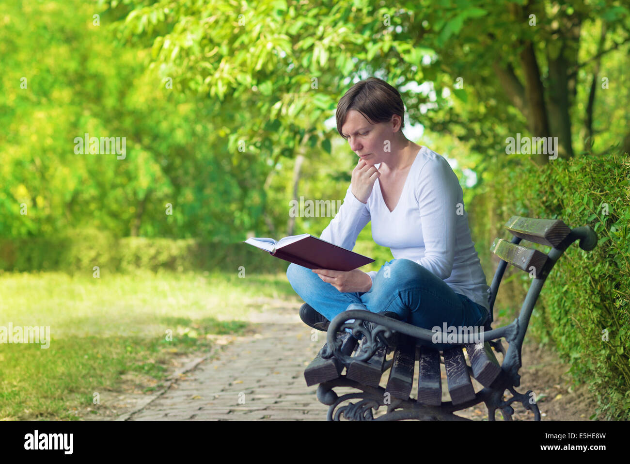 Schöne junge Frau sitzend auf einer Bank im Park an einem hellen Sommertag Hardcover-Buch zu lesen. Stockfoto