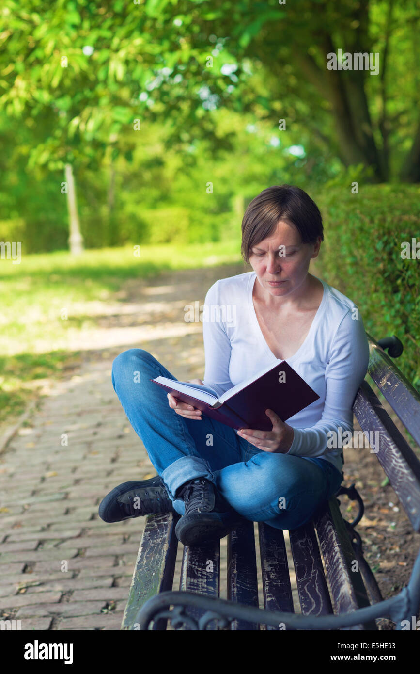 Schöne junge Frau sitzend auf einer Bank im Park an einem hellen Sommertag Hardcover-Buch zu lesen. Stockfoto