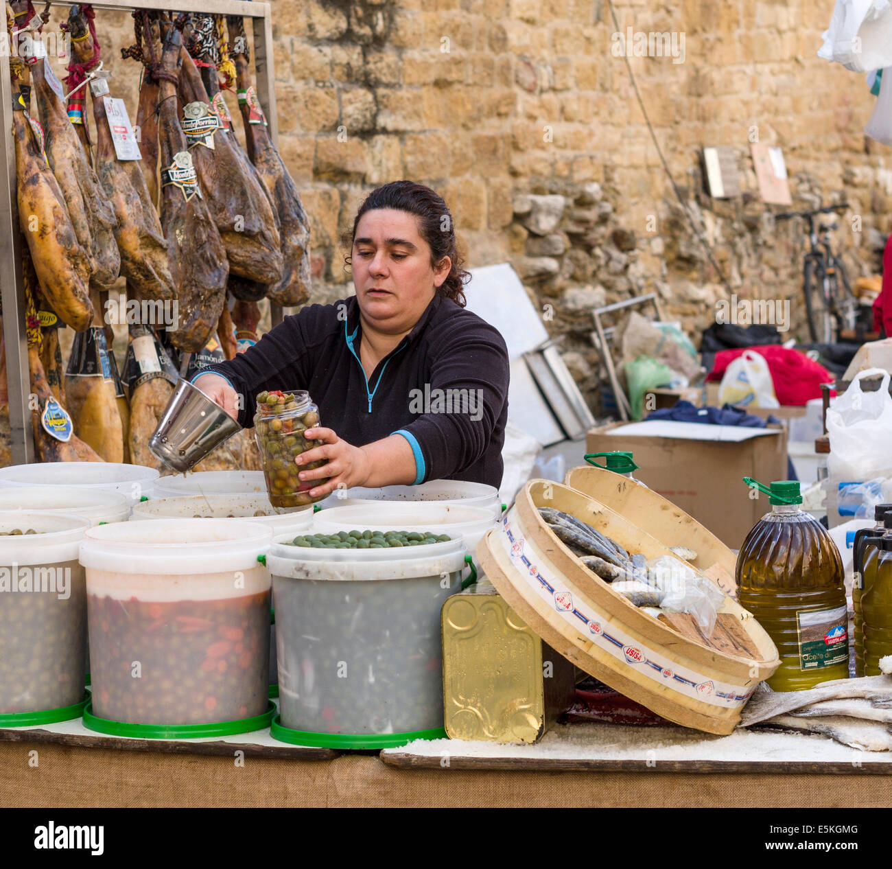 Messen Sie die Oliven. Eine Markt-Verkäufer schöpft aus einem Glas voller eingelegte Oliven. Schinken Fisch und Olivenöl sind im Hintergrund Stockfoto